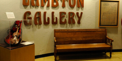 Violin under glass case and wooden bench under Lambton Gallery sign